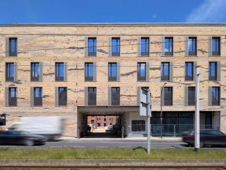 Eine moderne beige Steinfassade ist durchzogen von dunkelgrünen, grauen und roten Steinen. Vor dem Erdgeschoss stehen noch Bauzäune, die Straße vor dem Neubau ist vielbefahren.